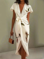 Close crop showing the ivory wrap dress bodice, tie-waist bow and flutter sleeve detail on a sage green backdrop