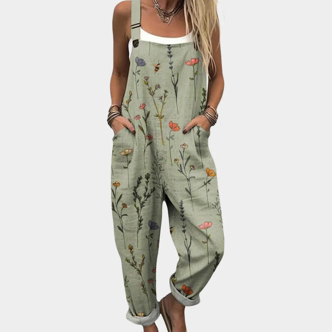 Woman wearing a sage green botanical print jumpsuit with wide straps, cuffed hems and sandals, hands in front pockets