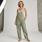 Full-length front view of a model in a green wildflower print overall-style jumpsuit styled with a white singlet and flat sandals