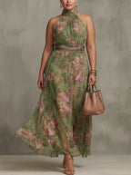 Woman modelling a green floral chiffon halter maxi dress with gathered bodice, matching waist sash and gold heeled sandals, holding a beige handbag