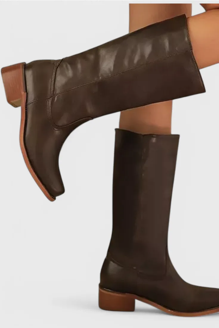 Close-up side profile of dark brown knee-high leather boots on a model, showing stacked heel and pointed toe shape