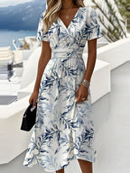 Woman wearing a white midi dress with navy leaf print, V-neckline and cinched waist, standing near a coastal terrace.