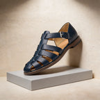 Navy leather fisherman sandal shown at an angle on a grey plinth, revealing the woven strap design and buckle closure.