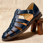 Close-up side view of navy leather fisherman sandal resting on a wooden stool, showing the stitched sole and tan leather lining.
