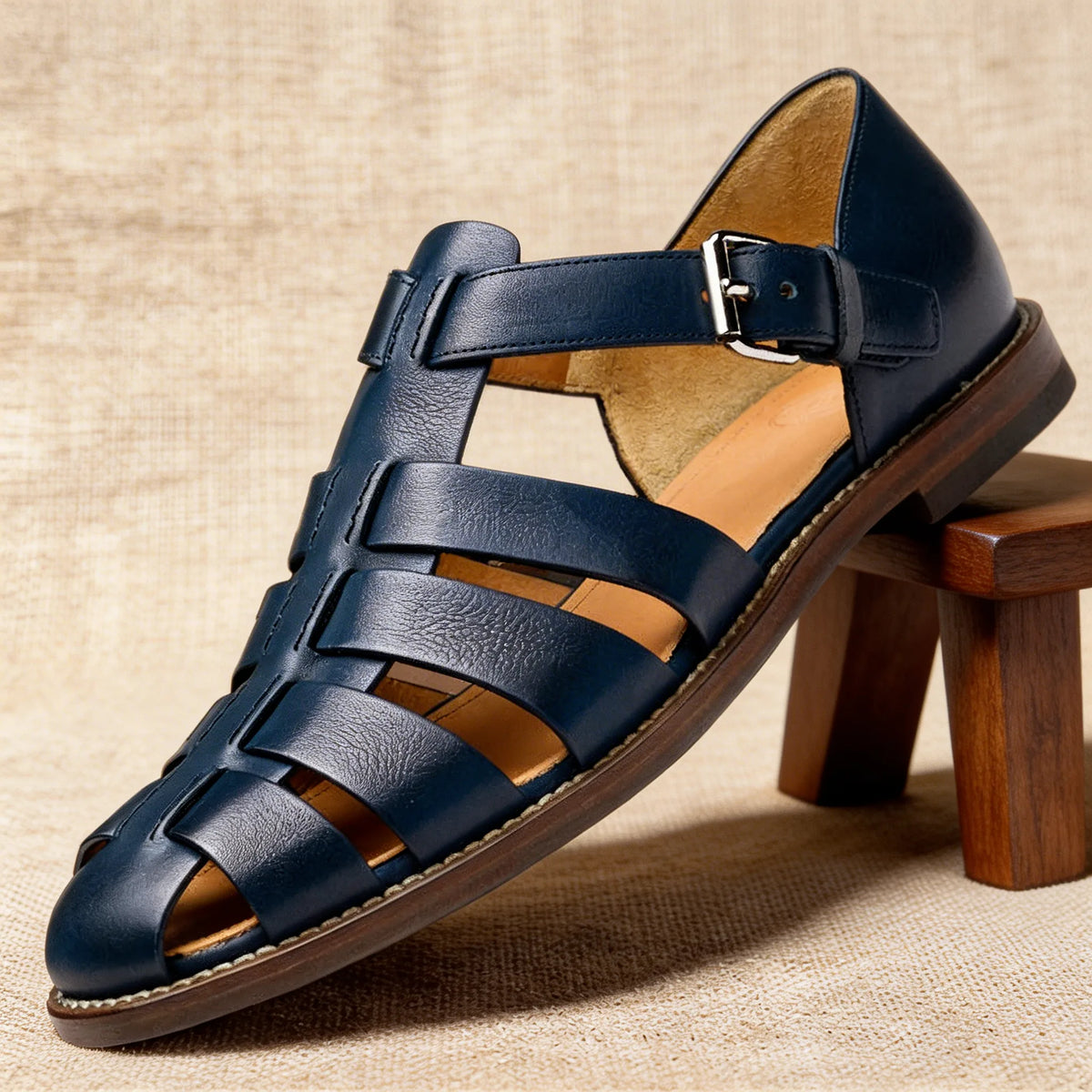 Close-up side view of navy leather fisherman sandal resting on a wooden stool, showing the stitched sole and tan leather lining.
