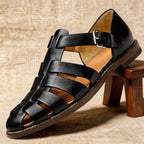 Black leather fisherman sandal with interwoven straps and silver buckle, displayed on a wooden stool against a woven backdrop.