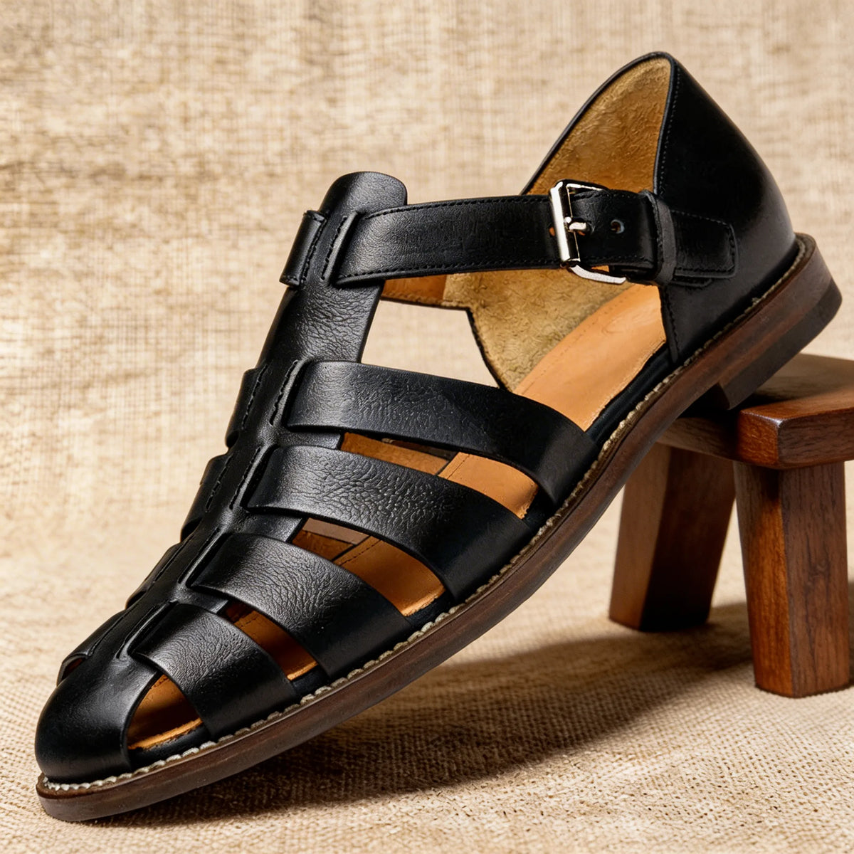 Black leather fisherman sandal with interwoven straps and silver buckle, displayed on a wooden stool against a woven backdrop.