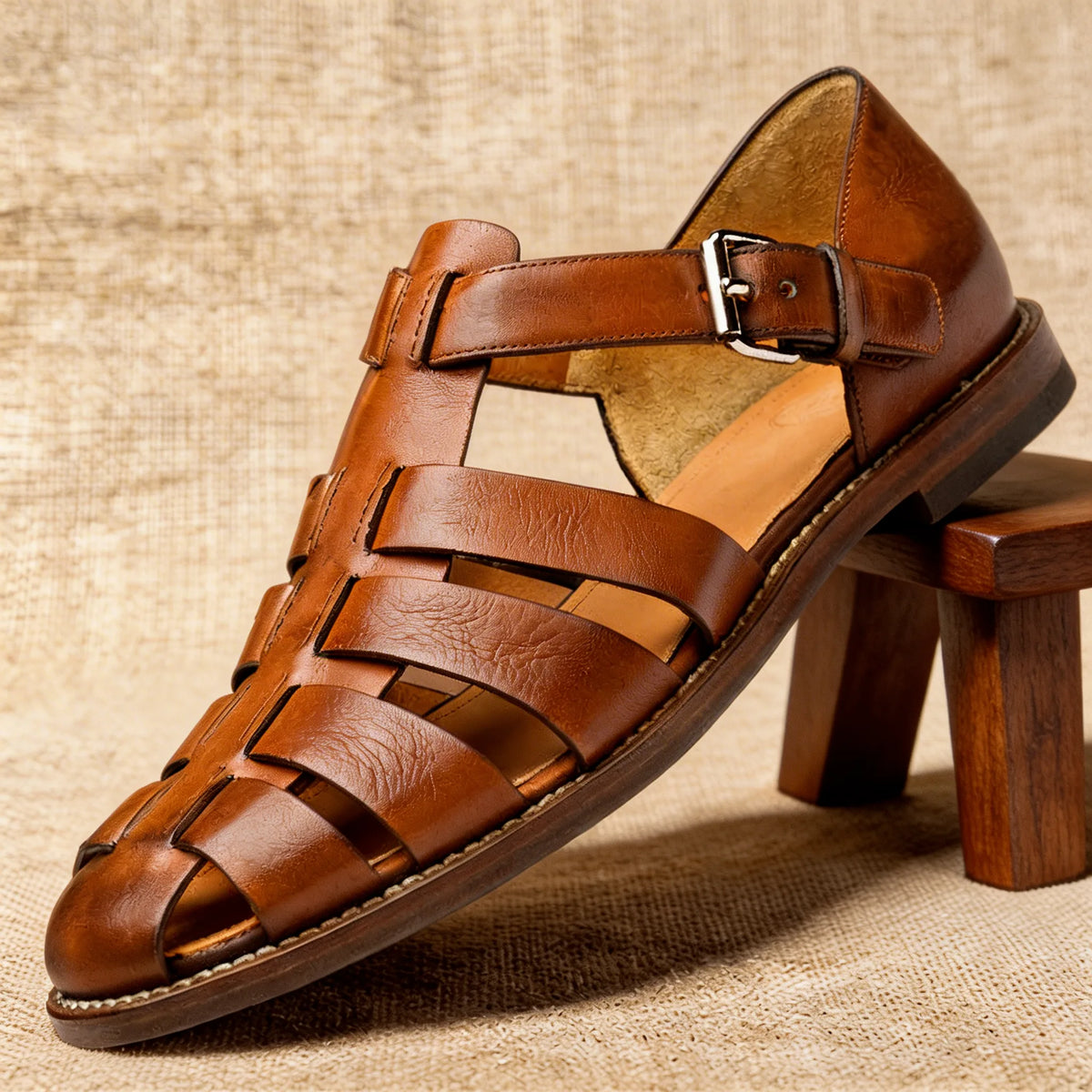 Light brown leather fisherman sandal with visible grain texture and tan insole, propped against a small wooden stool.