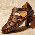 Dark brown leather fisherman sandal with rich tone and stitched trim, positioned on a wooden stool against a linen backdrop.