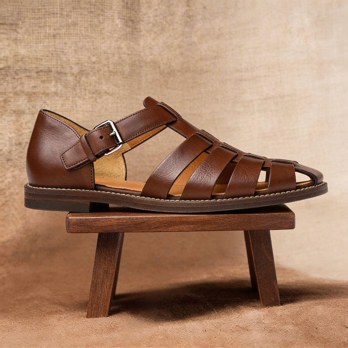 Dark brown leather fisherman sandal viewed in full profile on a wooden stool, showing the closed toe and low heel sole.