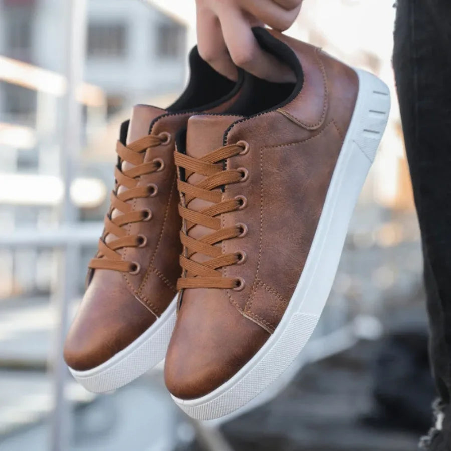 Hand holding a pair of brown leather sneakers by the heel tabs outdoors with laces crossed neatly