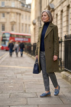 Woman walking down a city street in an olive coat and navy jumper, wearing dusty blue tasselled loafers