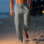 Man walking barefoot on the beach at dusk wearing light grey linen drawstring trousers with side pockets.