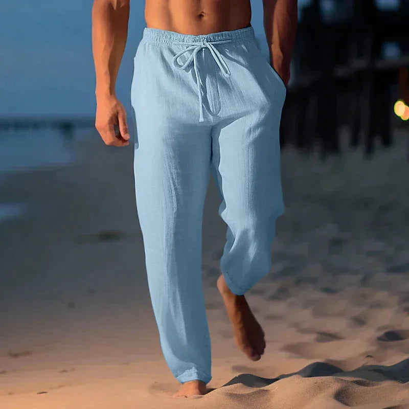 Front view of pale sky blue linen trousers with elasticated drawstring waist, worn on sand by the shore.