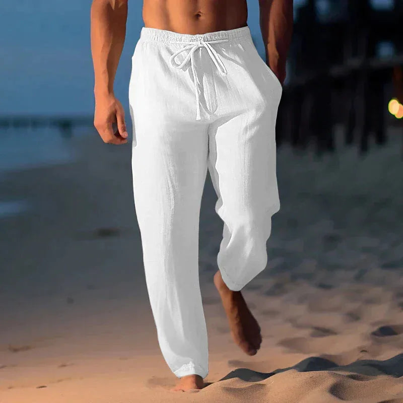 Close-up front shot of white linen trousers with tied drawstring and relaxed straight leg, beach background.