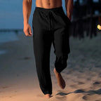 Black linen drawstring trousers shown in motion on sand, highlighting the loose breathable fit and ankle hem.