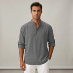 Front view of a man wearing a grey linen-blend popover shirt with a band collar and rolled-up long sleeves, paired with white pants.
