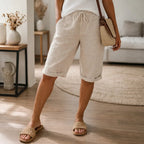 Cropped front view of cuffed beige linen Bermuda shorts with drawstring waist, styled with raffia slides and a crossbody bag