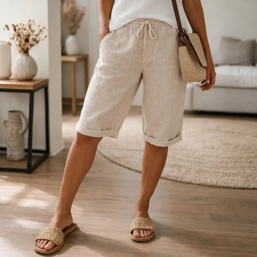 Cropped front view of cuffed beige linen Bermuda shorts with drawstring waist, styled with raffia slides and a crossbody bag