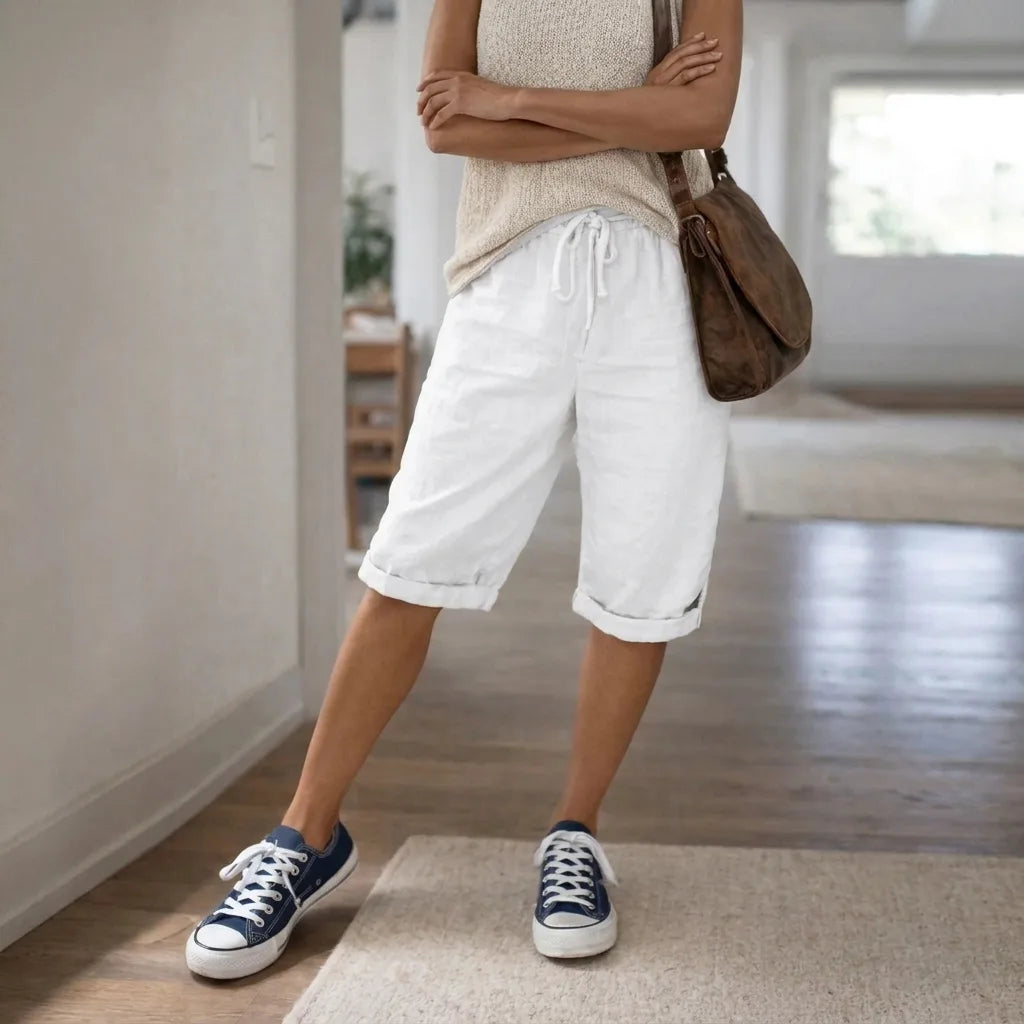 White linen Bermuda shorts with rolled cuffs and drawstring waist, styled casually with navy canvas sneakers