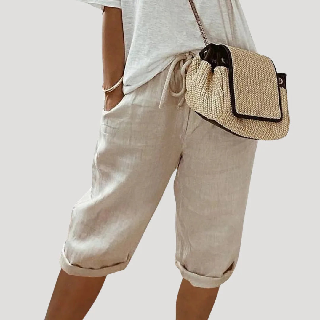 Model wearing beige linen bermuda shorts with elastic drawstring waist, cuffed hems and woven crossbody bag