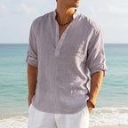 Man in a grey linen Henley shirt with rolled cuffs, paired with white trousers on the shore