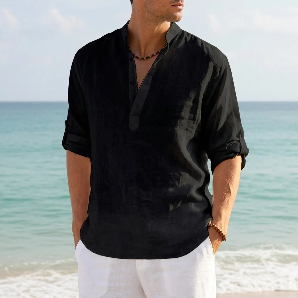 Man in a black linen Henley shirt with banded collar, hands in pockets on a sandy beach