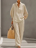Beige two-piece co-ord shown in full, loose batwing-sleeve top tucked lightly over flowing wide-leg trousers and a straw tote.