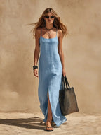 Model walks in a light blue linen slip maxi dress with scoop neck and thigh-high side split, carrying a woven tote bag