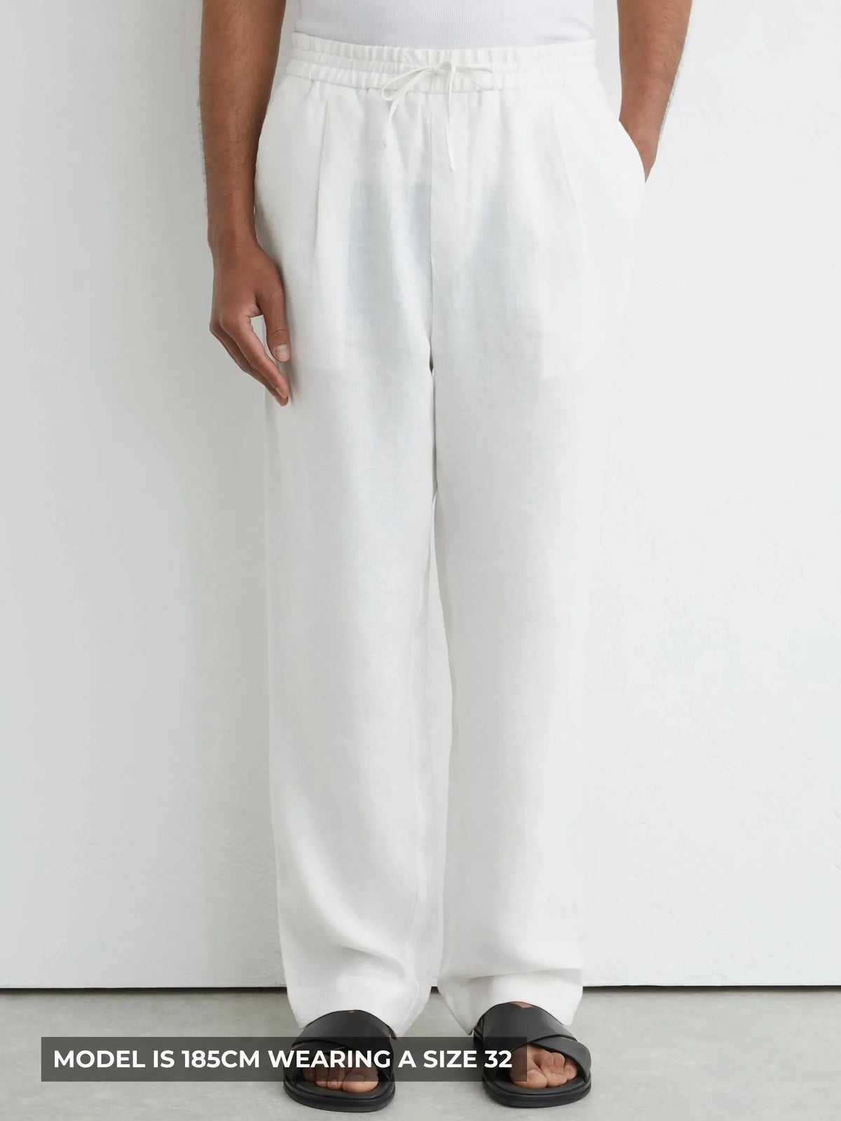 Front view of man wearing white relaxed-fit linen trousers with drawstring waist and black slide sandals