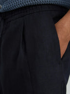 Close-up of hand in slanted pocket of navy linen trousers showing pleat detail at waistband
