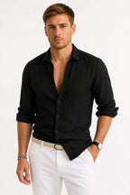 Man wearing a black linen shirt with classic collar, buttoned front and rolled-up sleeves, worn open at the neck with white pants.