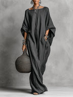 Front view of a woman in a dark grey loose cotton-linen maxi dress with batwing sleeves, styled with layered silver jewellery and a woven bag