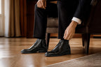 Man in a black suit adjusting his trousers while wearing black leather lace-up chukka boots indoors
