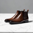 Pair of brown leather Chelsea boots with pointed toe and elastic side panels on marble surface
