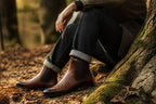 Man wearing brown leather Chelsea boots with cuffed jeans, seated outdoors near a tree