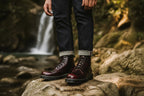 Man wearing burgundy leather combat boots with rolled denim jeans, standing on a rock near a waterfall.
