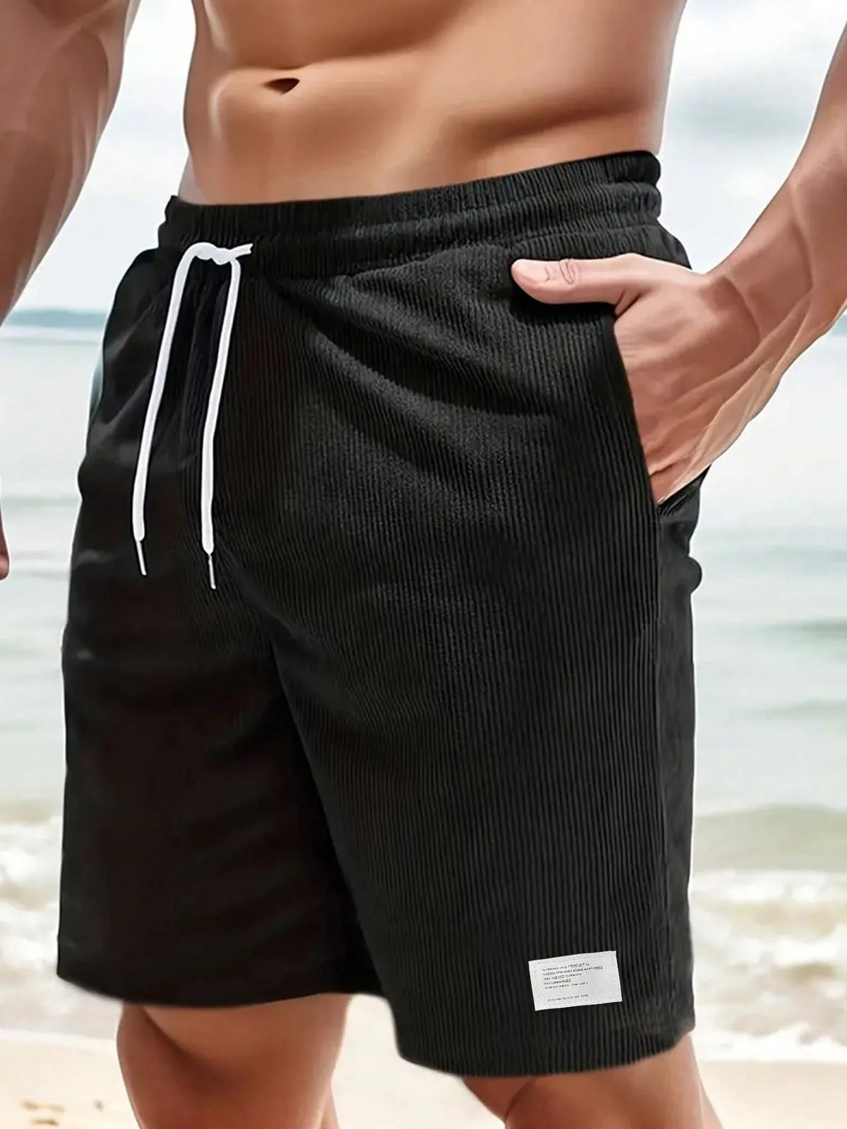 Close-up front view of black ribbed corduroy shorts with white drawstring, beach background