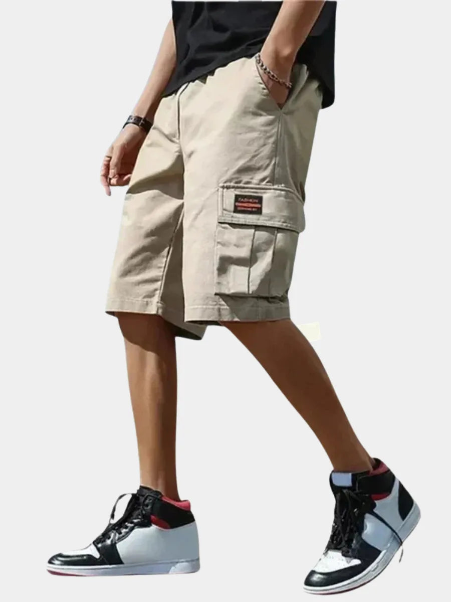Beige cotton cargo shorts shown from the side on a model, featuring a side cargo pocket with flap and fashion patch