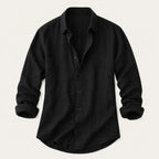 Flat lay of a black cotton-linen button-up shirt with rolled sleeves on a plain background