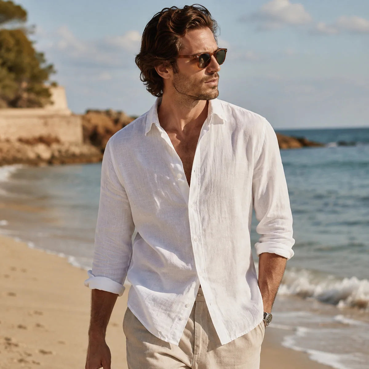 Man walking on a beach in an open white linen shirt and cream trousers near the shoreline