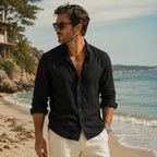 Man standing on a beach in a black linen button-up shirt with sleeves rolled, ocean in background