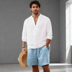 Man wearing a white linen popover shirt with light blue drawstring shorts, holding a straw hat, full outfit view