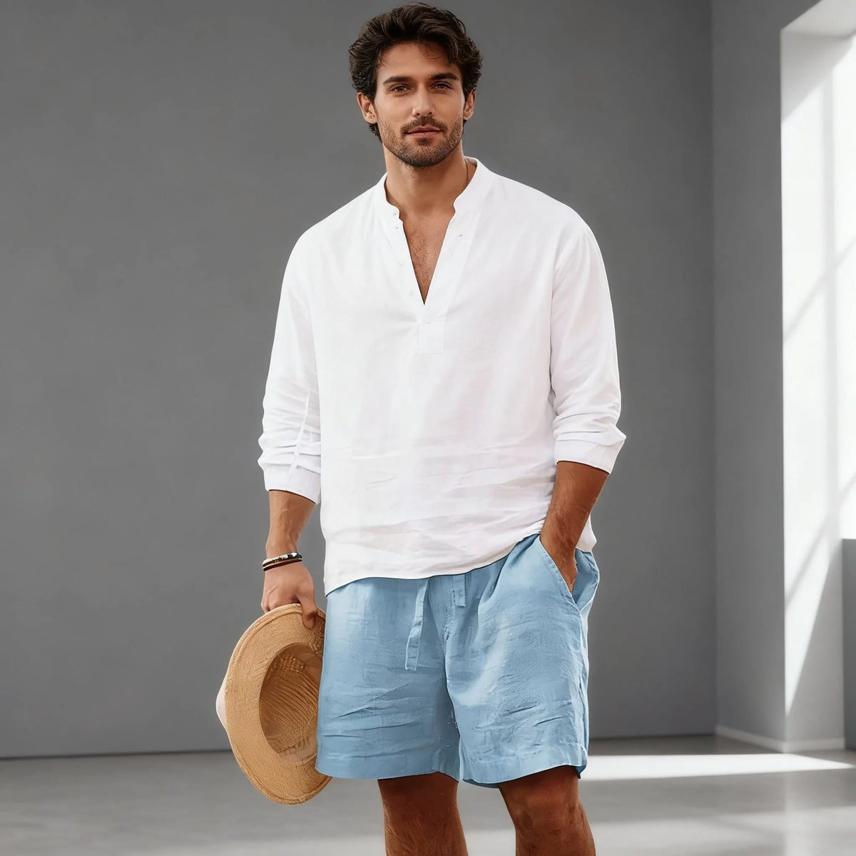 Man wearing a white linen popover shirt with light blue drawstring shorts, holding a straw hat, full outfit view