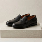 Side view of black leather penny loafers with hand-stitched moc toe and tan leather lining, shown on a stone pedestal.