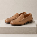 Pair of sand tan leather penny loafers with classic strap detail and stacked wooden heel, angled on a stone block.