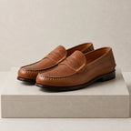 Tan brown leather penny loafers with visible hand stitching and leather sole, displayed on a textured stone surface.