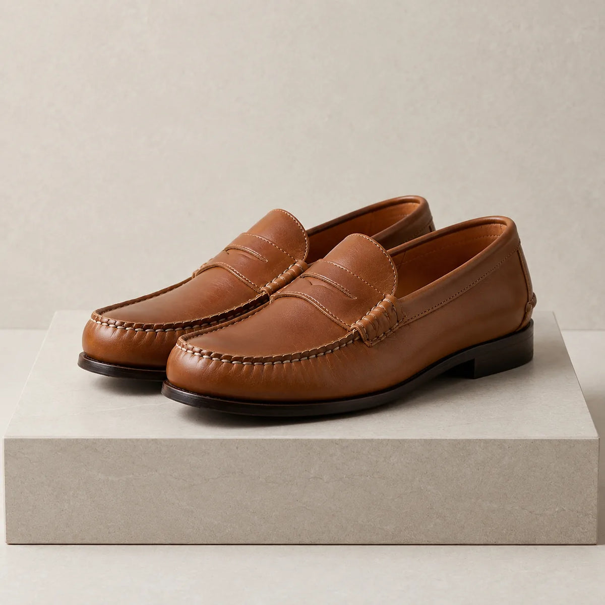 Tan brown leather penny loafers with visible hand stitching and leather sole, displayed on a textured stone surface.