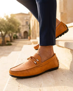 Tan leather driving loafer with woven bit buckle detail, worn with navy tailored pants on a sunlit stone staircase