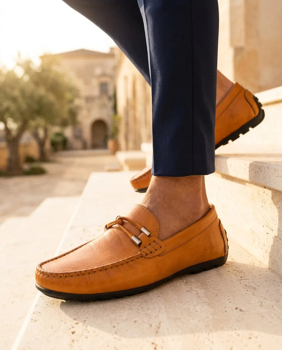 Tan leather driving loafer with woven bit buckle detail, worn with navy tailored pants on a sunlit stone staircase